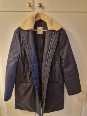 Mörkblå parkajacka från Boomerang Large - Parkajacka från Boomerang i mörkblått med mjuk teddykrage i beige. Jackan har både dragkedja och knappar framtill, två stora fickor och en rak, längre passform. Perfekt för kalla dagar med sin fodrade insida och stilrena look.