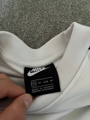 Vit Nike sweatshirt - Säljer en fräsch vit sweatshirt från Nike i storlek XS. Tröjan har svart Nike-logga på bröstet och svarta detaljer längs ärmarna. Materialet är mjukt och känns sportigt, perfekt för chill eller träning. Rund hals och långa ärmar ger en clean look.