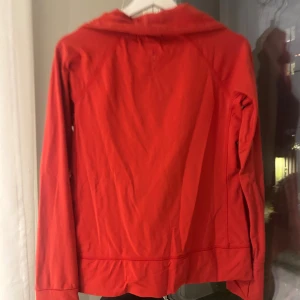 Röd vindjacka från New Apparel - Snygg röd vindjacka från New Apparel i storlek M. Jackan har dragkedja framtill, huva och diskret logga på bröstet. Materialet är lätt och känns som syntet, perfekt för blåsiga dagar. Enkel design med lång ärm och smidig passform.