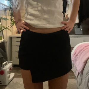 Lågmidjad minikjol - Svart kortkjol med snygg asymmetrisk nederkant. Kjolen har en enkel och stilren design som sitter midjehögt och är lätt att matcha. Perfekt för dig som gillar minimalistisk stil och vill ha något som sticker ut lite extra. Det är en ”skort”, alltså kombination av kjol och shorts.