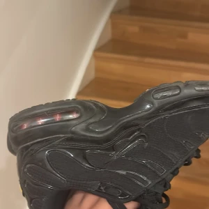 Nike Air Max Plus Triple Black - Säljer ett par Nike Air Max Plus i helsvart färg. Skorna har den ikoniska Tuned Air-sulan med synliga luftbubblor och vågformade detaljer på ovandelen. Materialmix av syntet och textil, med snörning och platt sula. Perfekta för dig som gillar streetwear och sportig stil.