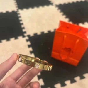 Guldigt armband med blommönster - Snyggt armband i guldig metall med blommönster i rosa och blått. Armbandet har en stilren, rund form och detaljerade kanter med räfflad struktur. Perfekt accessoar för att lyfta din outfit med en färgklick och trendig design.