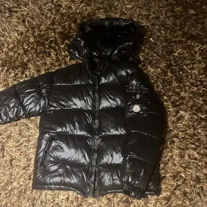 Svart Moncler pufferjacka med huva - Säljer en svart pufferjacka från Moncler med glansig finish och huva. Jackan har klassisk Moncler-logga på ärmen, två sidofickor med dragkedja och quiltad design. Perfekt för kalla dagar och har en riktigt snygg streetstil.