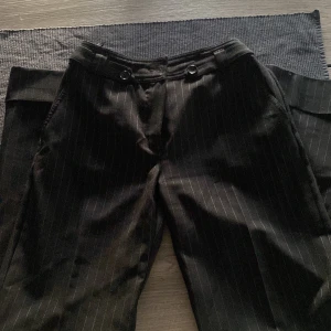 Svarta randiga kostymbyxor Lindex 36 - Svarta kostymbyxor från Lindex i storlek 36 med tunna vita ränder. Byxorna har raka ben, sidfickor och bakfickor samt snygga knappar i midjan. Perfekta för en stilren och trendig look.