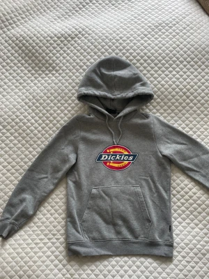 Ljusgrå  hoodie från Dickies XS - Ljusgrå hoodie från Dickies i storlek XS med klassisk logga i rött, gult och blått på bröstet. Tröjan har huva med snörning, magficka och är tillverkad i 96 % bomull, 4 % elestan. Perfekt för steetstyle.