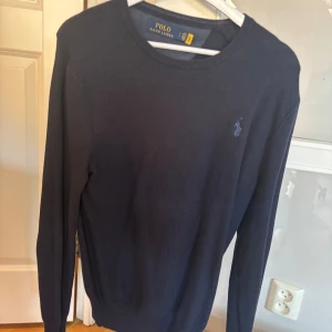 Marinblå stickad tröja Polo Ralph Lauren - Snygg marinblå stickad tröja från Polo Ralph Lauren med klassisk rund hals och diskret broderad logga på bröstet. Tröjan har långärmad passform och är perfekt för dig som gillar stilrena och tidlösa plagg. Passar dig som vill ha en enkel men schysst look.