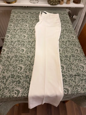 Vita låga kostymbyxor från BikBok, strl 38 - Snygga vita kostymbyxor från BikBok i rak och något utsvängd modell. Byxorna har låg midja, hällor för bälte, diskreta fickor bak och stängs med dragkedja och hake. Perfekta för en clean och stilren look. Helt nya, aldrig använda! Köpta för 499kr