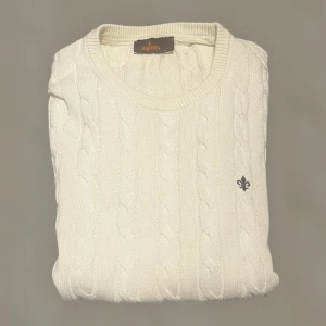 Vit kabelstickad tröja från Morris - Snygg vit kabelstickad tröja från Morris med rund hals och diskret logga broderad på bröstet. Tröjan har långärmad passform och är tillverkad i mjuk bomull, perfekt för dig som gillar klassisk och clean stil.