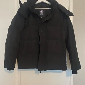 Svart pufferjacka från Canada Goose Man S/M - Svart pufferjacka från Canada Goose med huva och klassisk logga på ärmen. Jackan har dragkedja och knappar framtill, samt två stora fickor. Kvalitet som ny då den endast används fåtal gånger