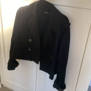Svart croppad trenchcoat från Gina Tricot - Svart croppad rock från Gina Tricot med klassisk krage och stora knappar framtill. Jackan har långa ärmar med snygga knappdetaljer vid mudden och en stilren, rak passform. Perfekt för dig som gillar minimalistisk och trendig stil.