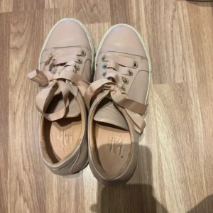 Beige sneakers från Billie - Snygga beige sneakers från Billie med breda satinliknande snören och guldfärgade öljetter. Skorna har rund tå och platt sula, tillverkade i skinn med en stilren design. Perfekta för dig som gillar neutrala färger och vill ha en clean look.