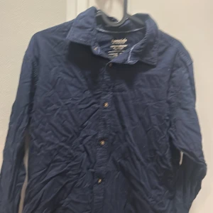 Mörkblå skjorta från Esprit - Snygg mörkblå skjorta från Esprit med klassisk krage och knappar framtill. Skjortan har lång ärm och är gjord i mjuk bomull som känns skön mot huden. Perfekt för dig som gillar stilrena och enkla plagg med en chill vibe.