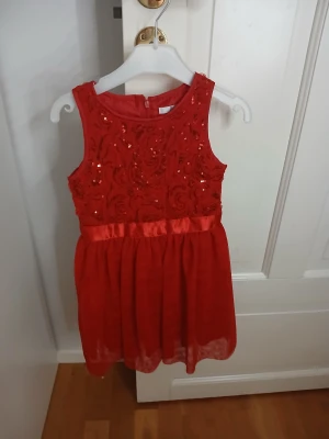 Röd glittrig klänning från Åhléns - Superfin röd klänning från Åhléns i storlek 104. Klänningen har glittriga paljetter och broderade detaljer på överdelen, ett satinband i midjan och ett skirt, lätt plisserat kjol parti. Ärmlös modell med dragkedja i ryggen, perfekt för speciella tillfällen.