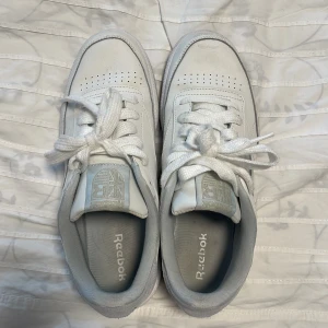 Vita Reebok sneakers med snörning - Fräscha vita Reebok sneakers med klassisk låg profil och rund tå. Skorna har snörning, perforerade detaljer på tån och Reebok-logga i silver på sidan och hälen. Ovandelen är i skinn med mockadetaljer bak, och innersulan är mjuk för extra komfort.