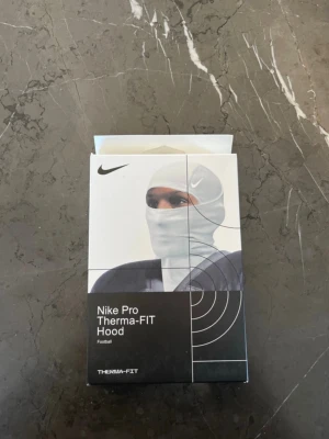Nike Pro Therma-FIT Hood - Nike Pro Therma-FIT Hood är en svart, tight och sportig huva designad för fotboll. Den är tillverkad i ett värmande syntetmaterial som håller dig varm under träning eller match. Perfekt för kalla dagar på planen.