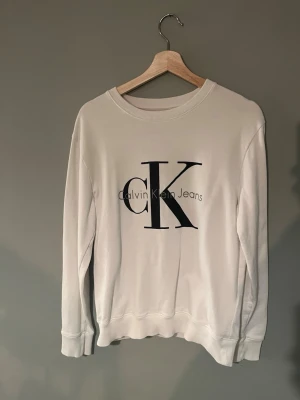 Vit sweatshirt från Calvin Klein Jeans - Säljer en stilren vit sweatshirt från Calvin Klein Jeans med stor svart CK-logga framtill. Tröjan har rund halsringning, långa ärmar och ribbade muddar vid ärmslut och nederkant. Perfekt för en clean och avslappnad look.