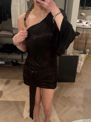 Svart glittrig oneshoulder klänning - Säljer en svart glittrig kortklänning med oneshoulder-design och lång, vid ärm på ena sidan. Klänningen har snygg drapering och knyt i midjan som ger en cool look. 