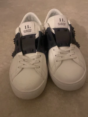 Valentino Rockstud vita sneakers - Säljer ett par vita Valentino Rockstud Untitled sneakers i skinn med svarta detaljer och ikoniska nitar över vristen. Klassisk rund tå, snörning och platt sula. Märket och modellen står tryckt på plösen. Snygg kontrast mellan vitt och svart, perfekt för dig som gillar statement-detaljer.