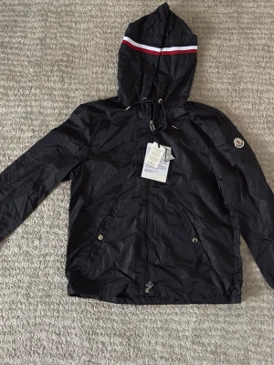 Svart vindjacka från Moncler - Snygg svart vindjacka från Moncler med huva och dragkedja framtill. Jackan har en vit och röd rand på huvan, snörning vid halsen och två fickor med tryckknappar. Perfekt för dig som vill ha en stilren och sportig look.