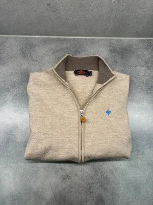 Morris merinoull  - Snygg beige kofta från Morris i 100% merinoull. Modellen har hel dragkedja framtill, ribbad krage och muddar samt en blå broderad detalj på bröstet. Perfekt för lager-på-lager och chillare dagar.