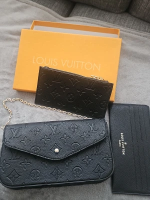 Svart axelväska med kedja - Säljer en svart axelväska  med klassiskt monogrammönster i skinn. Väskan har en snygg guldfärgad kedja och medföljande plånbok och korthållare i samma stil. Perfekt för dig som gillar lyxiga detaljer och ikonisk design.