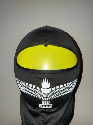 Svart balaclava med vit logga - Säljer en svart balaclava med vit grafisk logga framtill och stjärndetaljer. Materialet är stretchigt och syntetiskt, perfekt för att täcka hela ansiktet. Passformen är tight och designen är enkel men ändå unik med trycket framtill. Äkta använd i musik video