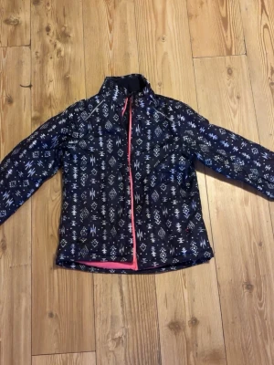 Mönstrad vindjacka från Crivit - Säljer en mörkblå vindjacka från Crivit med coolt geometriskt mönster i vitt och grått. Jackan har rosa dragkedja och insida, samt hög krage och långa ärmar. Perfekt för dig som gillar att sticka ut med din stil.