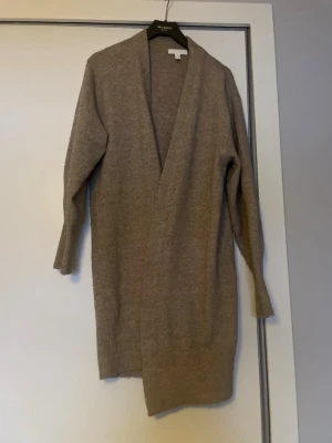 HM Beige lång kofta i ullblandning - HM Mysig beige lång kofta från John Baner. Koftan har en öppen front utan knappar, ribbade muddar och är tillverkad i mjuk ullblandning. Perfekt att slänga över en t-shirt eller hoodie när det blir kyligt ute.ca 120 cm2 från hals 