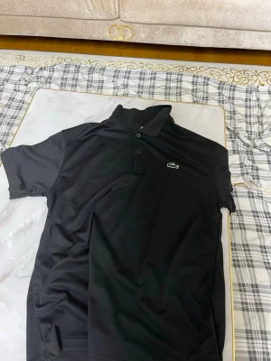 Svart pikétröja från Lacoste - Klassisk svart pikétröja från Lacoste med den ikoniska krokodilloggan broderad på bröstet. Tröjan har korta ärmar, krage och knäppning med tre knappar framtill. Tillverkad i bomull för en skön och luftig känsla.