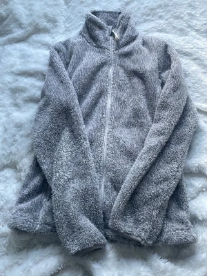 Grå teddyjacka med dragkedja - Mysig grå teddyjacka med hög krage och hel dragkedja framtill. Jackan är fluffig och har långärmad design, perfekt för kyliga dagar. Passar dig som gillar en avslappnad och bekväm stil.