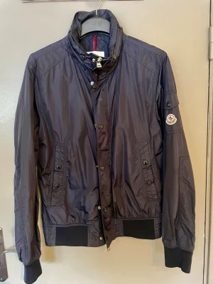 Mörkblå vindjacka från Moncler - Säljer en mörkblå vindjacka från Moncler med hög krage, knapp- och dragkedjestängning samt ribbade muddar. Jackan har Moncler-logga på ärmen, flera fickor med knappar och är tillverkad i lätt polyester. Perfekt för dig som vill ha en clean och stilren look.