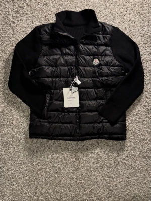 Moncler Cardigan - Svart Cardigan från Moncler i storlek L. Jackan har quiltad front i blank polyester och stickade ärmar samt hög krage. Klassisk Moncler-logga på bröstet och dragkedja framtill. Perfekt för dig som vill ha en clean och stilren look. Nypris ligger på 10000kr