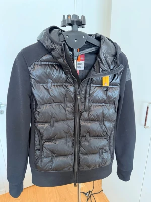 Svart cardigan från Parajumpers - Snygg svart cardigan från Parajumpers med quiltad front och huva. Jackan har dragkedja, två fickor fram och en gul detalj vid halsen. Perfekt för kyliga dagar och har en modern, sportig look.