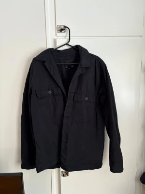 Svart overshirt från H&M - Säljer en svart overshirt från H&M med klassisk krage och två bröstfickor med knappar. Jackan är tillverkad i bomull och har långa ärmar samt knäppning framtill. Perfekt att slänga över en hoodie eller t-shirt.