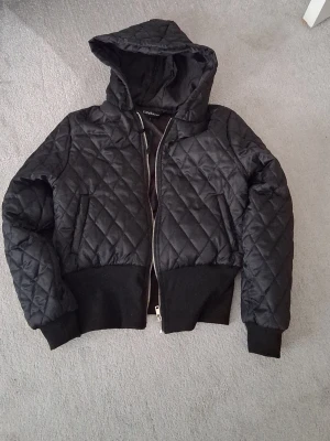 Svart quiltad bomberjacka EillyBazar - Säljer en svart quiltad bomberjacka från EillyBazar i storlek L. Jackan har huva, dragkedja framtill och breda muddar vid ärmar och midja. Perfekt för höst och vinter när du vill ha en stilren och bekväm look.