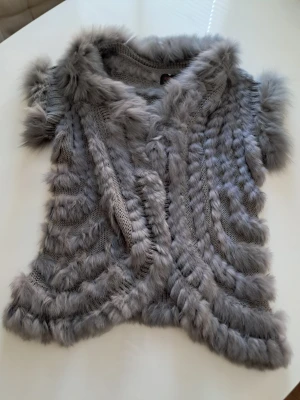 Grå pälsväst från Angelique.L M/L - Säljer en fluffig grå pälsväst från Angelique.L i storlek M/L. Västen har korta ärmar och är gjord av 70% kanin och 30% murmeldjur, med stickad bas och mjuka pälsdetaljer i ränder över hela plagget. Perfekt för dig som vill ha något extra mysigt och snyggt.
