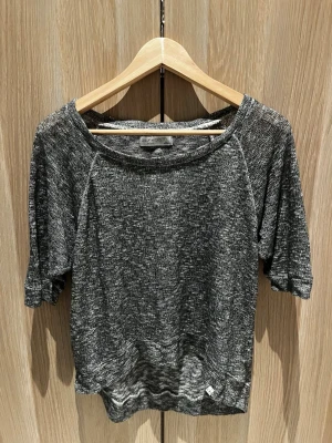 Melerad topp från Gilly Hicks - En gråmelerad topp från Gilly Hicks med avslappnad passform och korta ärmar. Materialet är mjukt och stickat, vilket ger en chill vibe. Rund halsringning och liten GH-logga vid nederkanten. Perfekt till jeans eller shorts.