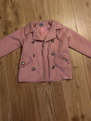 Rosa kappa från Kids by Voga - Säljer en rosa kappa från Kids by Voga i storlek 122-128. Jackan har dubbelknäppning med snygga metallknappar, bred krage och två fickor fram. Utsidan är mjuk och insidan har ett glansigt foder i matchande rosa ton. Perfekt för dig som vill ha en stilren och klassisk look. 