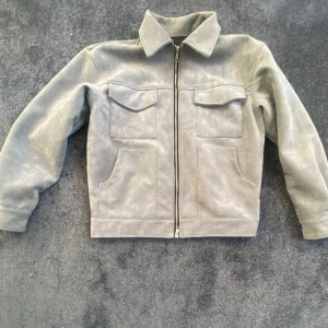 Grå mockajacka från HESNTLS - Säljer en stilren grå mockajacka från HESNTLS med två bröstfickor med lock och två sidofickor. Jackan har klassisk krage, raka ärmar och hel dragkedja framtill. Perfekt för dig som vill ha en clean look med lite edge.