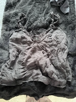 Svart  linnetopp från BikBok - Säljer en svart linnetopp från BikBok i storlek M. Toppen har smala axelband med volangdetaljer och en lätt rynkad, luftig passform. Materialet är tunt och känns som bomull, perfekt för varma dagar. Söt volangkant upptill och smockad rygg för extra komfort.