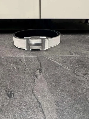 Vit och svart skärp med H-spänne - Säljer ett stilrent skärp i vitt och svart med ett stort metalliskt H-format spänne. Skärpet är gjort i präglat material som liknar skinn och har en clean look som passar till många outfits. Perfekt accessoar för dig som gillar statement-detaljer.