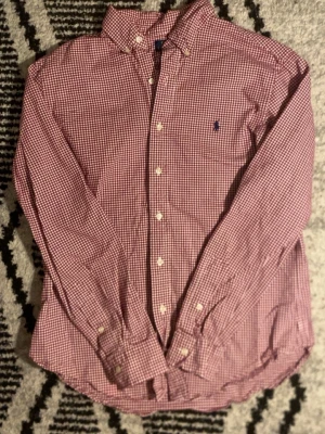  Polo Ralph Lauren skjorta  - Säljer en röd och vit rutig skjorta från Polo Ralph Lauren i slim fit. Skjortan har klassisk krage, knappar framtill och det ikoniska blå broderade logot på bröstet. Tillverkad i bomull med stretch för extra komfort.