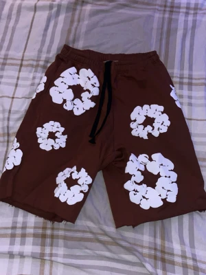 Bruna blommiga shorts från Denim Tears - Bruna shorts från Denim Tears med vita blommönster över hela plagget. De har elastisk midja med svart snörning och råa kanter nertill. 