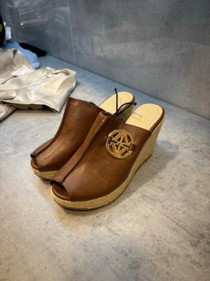 Bruna wedgesandaler med flätad sula - Snygga bruna sandaler med kilklack och öppen tå. Ovandelen är i skinnimitation och har en dekorativ guldfärgad metallogo på sidan. Sulan är flätad i naturmaterial för en somrig känsla. Perfekta till jeans eller klänning.