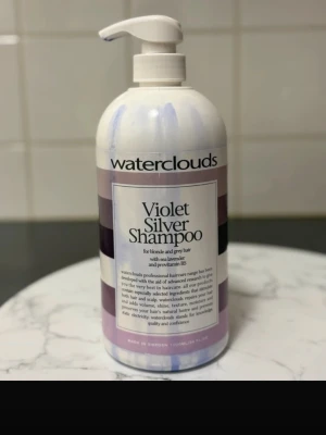 Waterclouds Violet Silver Shampoo - Stor pumpflaska med Waterclouds Violet Silver Shampoo, perfekt för blont eller grått hår. Innehåller havslavendel och provitamin B5. Schampot är djupt violettblått och hjälper till att neutralisera gula toner i håret. Flaskan rymmer 1000 ml och är vit med lila detaljer.