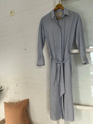 Randig skjortklänning Scotch & Soda XS - Säljer en långärmad skjortklänning från Scotch & Soda i storlek XS. Klänningen är vit med blå smala ränder, har klassisk krage, knytskärp i midjan och knappar framtill. Perfekt för en chill och stilren look.