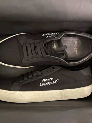 Svarta Saint Laurent sneakers - Snygga svarta sneakers från Saint Laurent med vit logotyp broderad på sidan. Skorna har rund tå, platt sula i beige och klassisk snörning. Ovandelen är i textil och detaljer i skinn. Perfekt för dig som gillar stilrena och exklusiva sneakers. PRIS KAN DISKUTERAS‼️‼️
