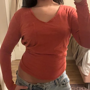 Långärmad tröja  - Säljer en röd långärmad topp från Superdry Studios i storlek EU 36. Toppen har v-ringad hals, en liten bröstficka och är figurnära i passformen. Materialet är mjukt och lätt, perfekt för dig som gillar enkel och stilren design.