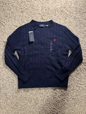 Stickad Tröja Ralph Lauren - Snygg marinblå stickad tröja från Polo Ralph Lauren i storlek M. Tröjan har kabelstickat mönster, rund hals och lång ärm. På bröstet finns den klassiska röda broderade loggan. Perfekt för dig som gillar stilrena och tidlösa plagg. Nypris ligger på 2100kr
