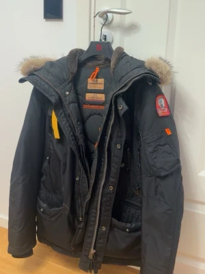 Svart Parajumpers parkajacka med päls - Svart parkajacka från Parajumpers med fluffig päls på huvan, coola detaljer som gul rem och patch på ärmen. Jackan har flera fickor, quiltad insida och dragkedja framtill. Perfekt för kalla dagar och har en riktigt snygg streetstil.